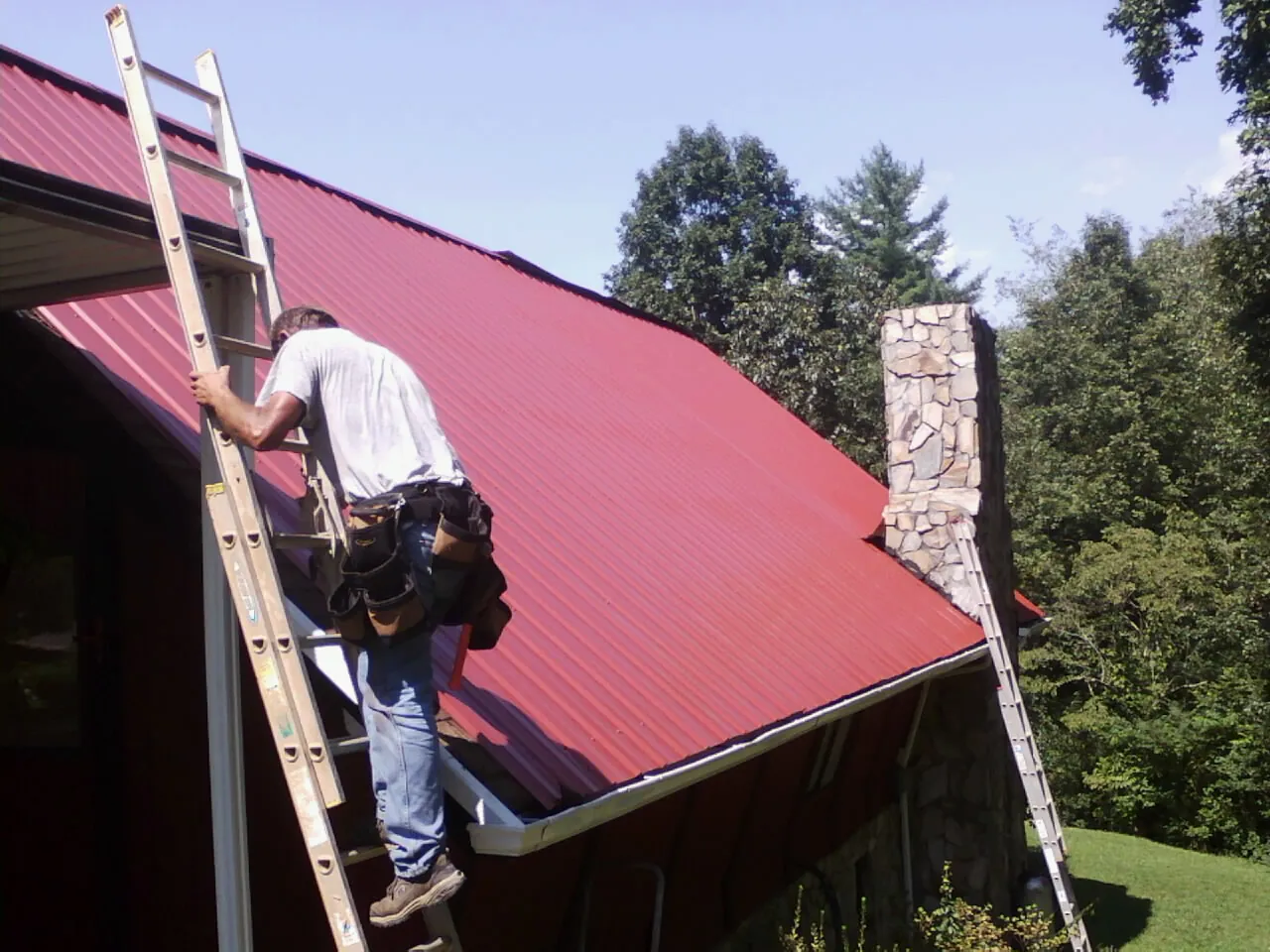 Roofer on a ladder beside a red metal roof and stone chimney — Soffit & Fascia Repair by Roofing Brothers in Rhodes